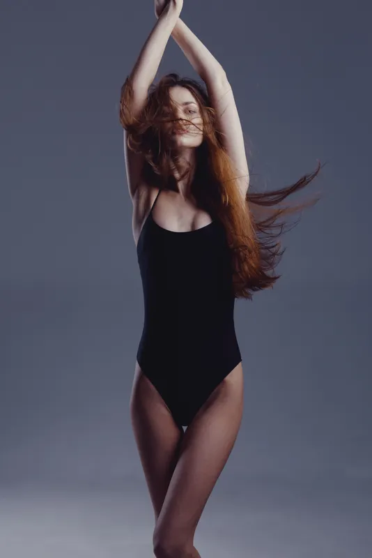 slender woman in black bodysuit dancing with arms raised against gray background