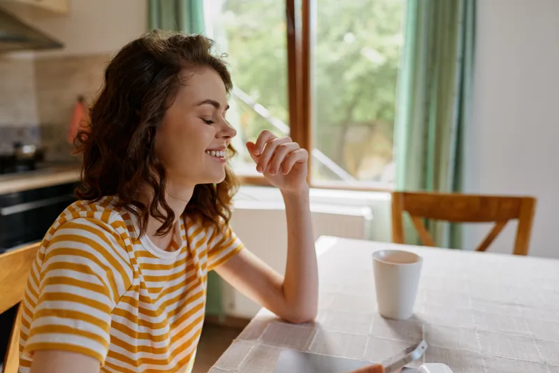 Smiling woman with wavy hair wearing a striped t shirt sits at a bright kitchen table enjoying a drink in a cozy home setting. Casual lifestyle, wellness, and everyday self care moment.
