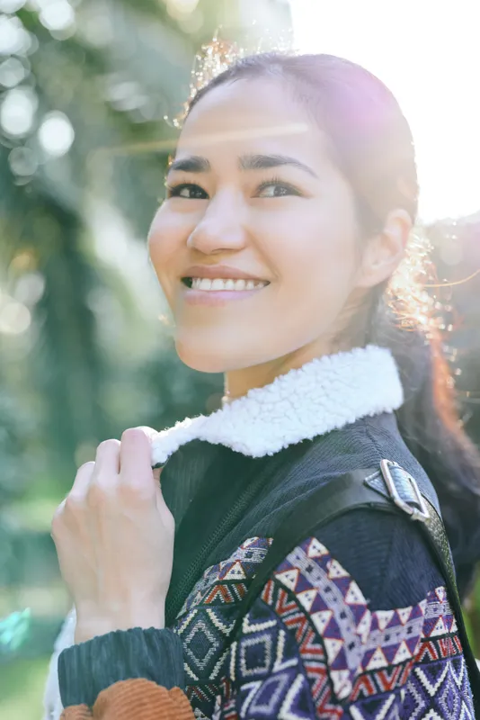 Woman with a warm smile in an outdoor setting wearing a patterned sweater and jacket, bright sunlight through trees, casual fashion and leisure stock photo for lifestyle use