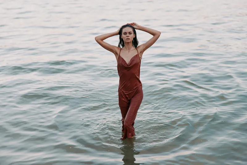 Portrait of a woman standing in shallow water, wearing a burgundy jumpsuit, hands raised behind head, calm expression, soft waves and warm light creating an intimate beach moment.
