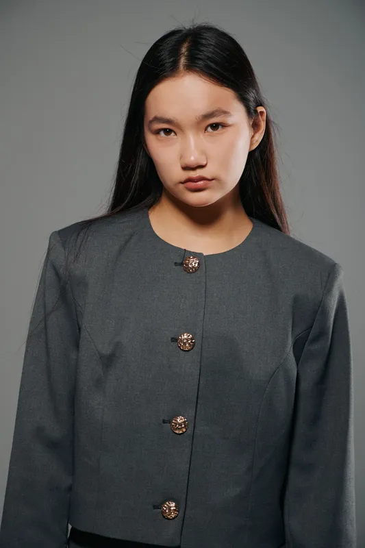 Woman portrait with long dark hair wearing dark gray jacket with decorative buttons standing against gray background. Serious expression, straight posture, studio lighting and simple fashion look.