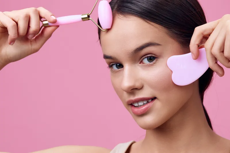 Young woman enjoying a skincare routine with a pink facial roller and gua sha tool against a vibrant pink background, conveying beauty and wellness