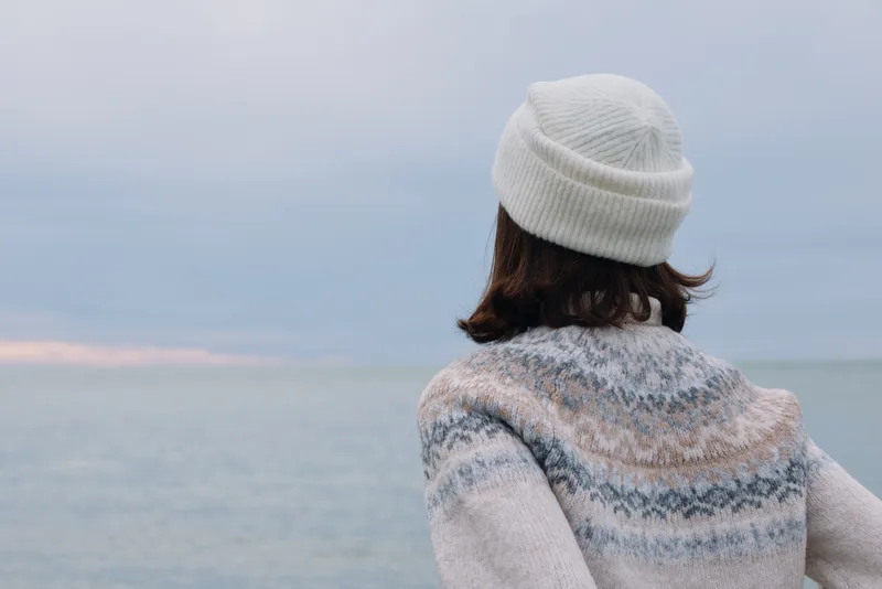 Person wearing a knitted sweater and white beanie, seen from behind, gazing at the calm ocean and distant horizon, creating a peaceful outdoor mood for travel or lifestyle projects