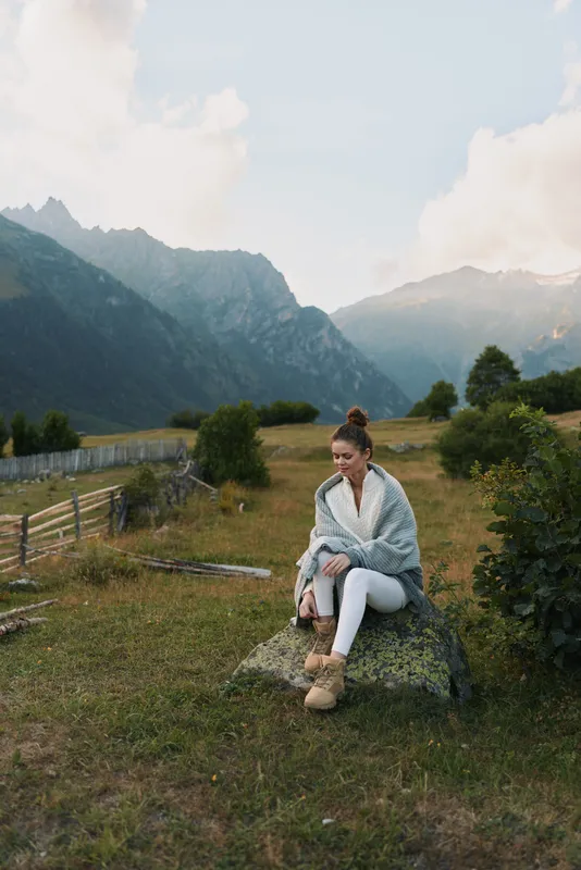A woman sits on a rock in a scenic meadow with distant mountains, conveying relaxed outdoors lifestyle and peaceful solitude in nature with light breeze, and open sky.