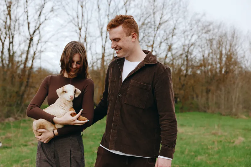 Young couple enjoys a moment outdoors as the woman holds a baby goat while the man smiles, both dressed in warm casual clothes during an autumn day in nature.