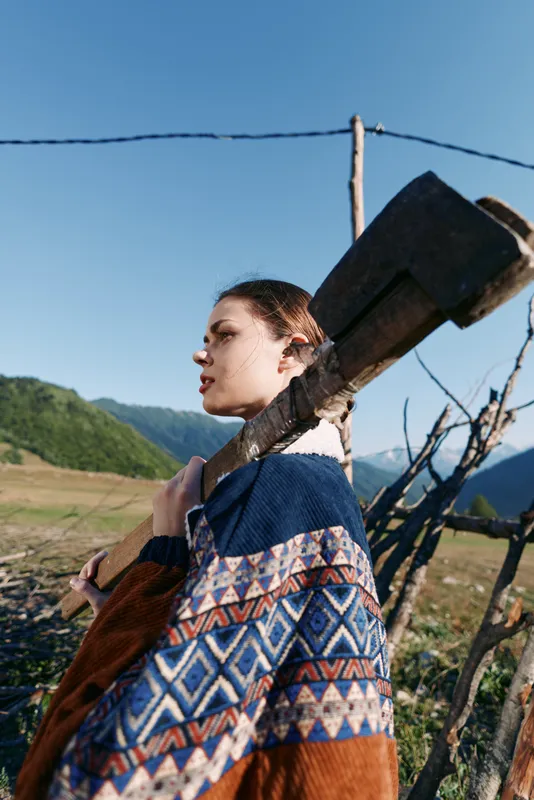woman ax countryside sweater fence mountains rural portrait of a young female carrying an ax on her shoulder in a rustic field, patterned sweater by wooden fence with clear blue sky