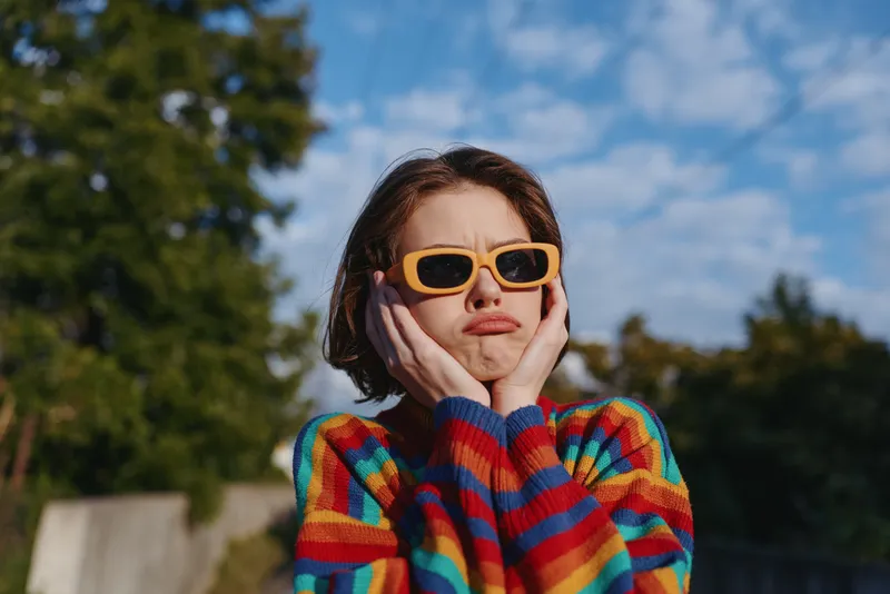 woman striped sweater sunglasses pout outdoor sunny young adult around 20s, short brown hair in bob, holding her cheeks and making a playful pout in a colorful casual outfit in park under blue sky