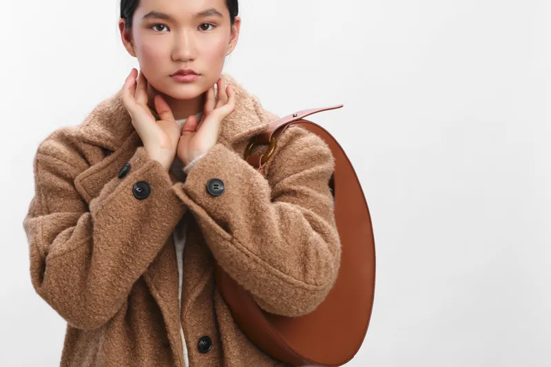 asian woman wearing a warm tan coat and carrying a round bag poses in a studio, showcasing contemporary fashion and a poised, confident attitude.