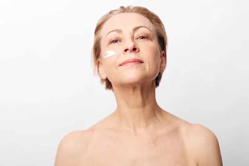 Confident mature woman applies white face cream to cheek, posing with bare shoulders on white background. Concept of skincare, aging, self-care, and natural beauty for older adults.