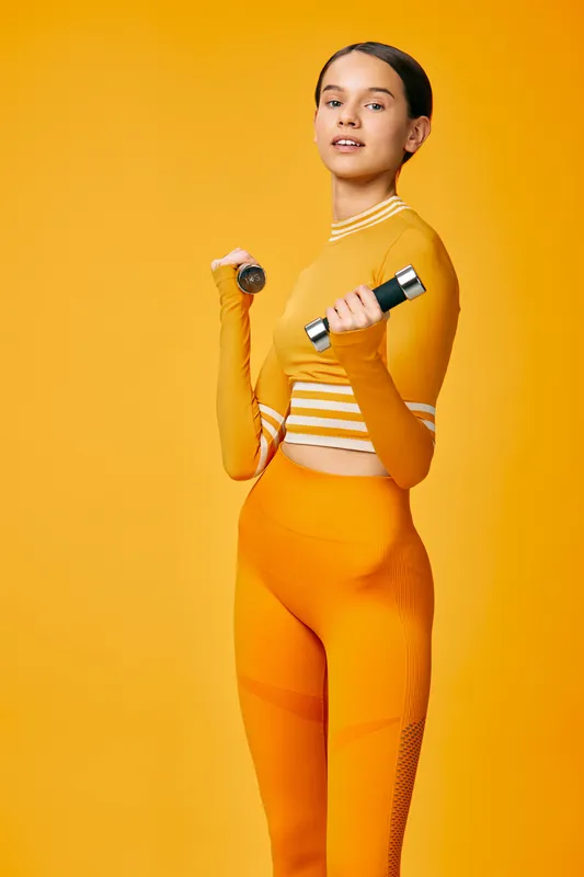 Young woman fitness portrait lifting small dumbbells in coordinated orange activewear, confident pose and engaged muscles, studio sports concept with seamless warm yellow background lighting.