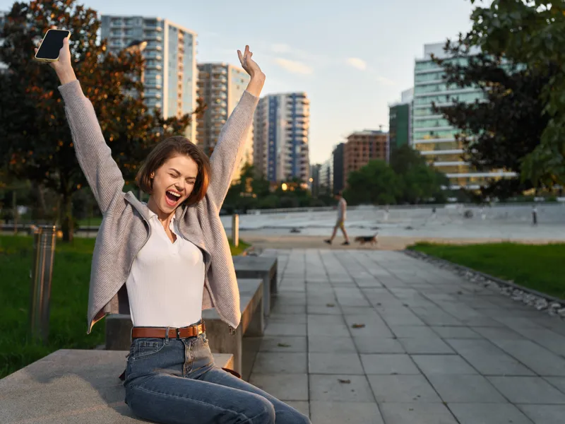 A cheerful young woman is seen outdoors in a modern city park. She smiles broadly, raises her arms in triumph, and holds a smartphone, conveying a lively, energetic urban mood.