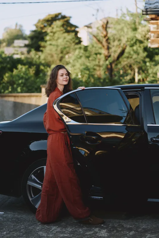 Woman in a red dress steps toward a sleek car, a candid lifestyle moment captured outdoors with an urban backdrop and confident, elegant mood