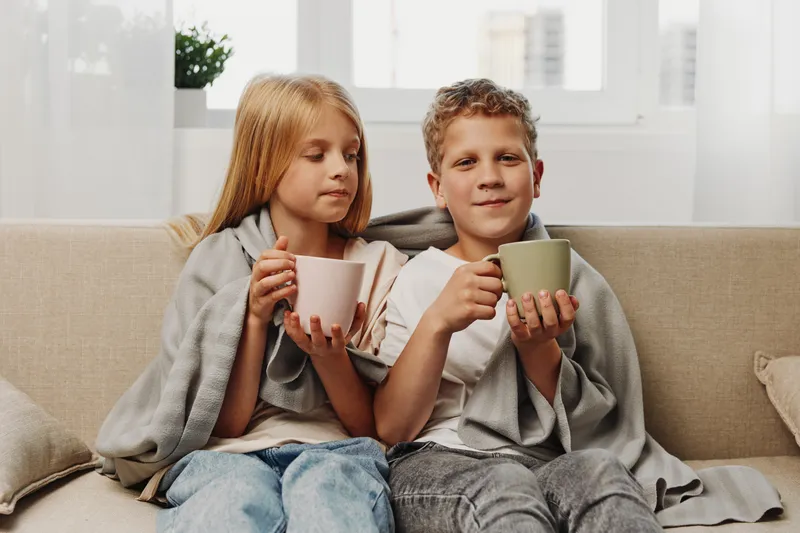 Two children wrapped in gray blankets hold ceramic mugs, sitting on a beige couch in a bright room by the window. Cozy atmosphere of relaxation, warmth, and family moments.