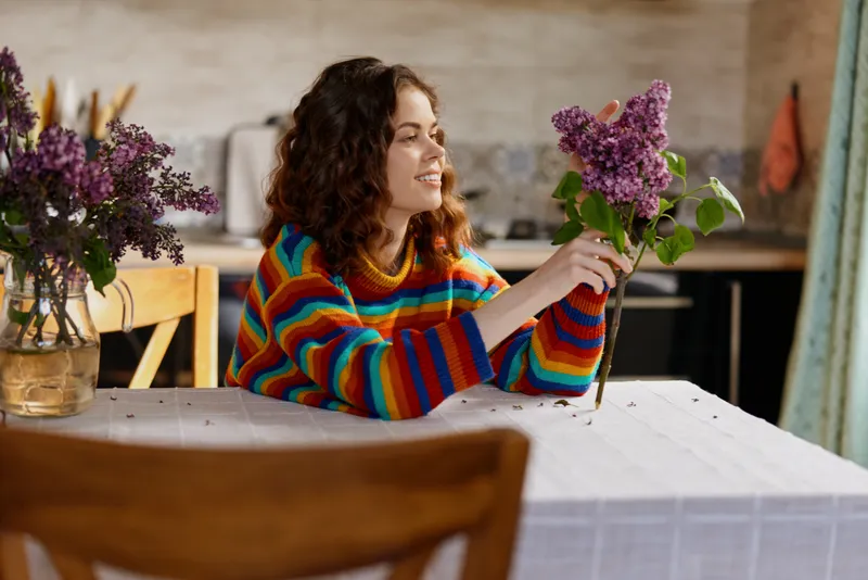 Young woman with curly brown hair wearing a rainbow striped sweater smiles while admiring a bouquet of lilac flowers resting on a white dining table in a cozy home setting