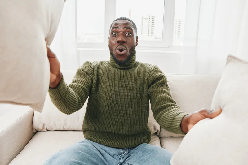 Excited Black man with pillows enjoying cozy indoor moments in a green sweater, expressing joy and surprise in a bright living room setting
