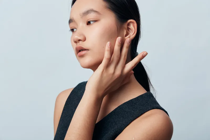 Portrait of a young woman with natural skin and minimal makeup touching her face, looking sideways on a light background. Beauty, fashion, and skincare concept with calm expression.