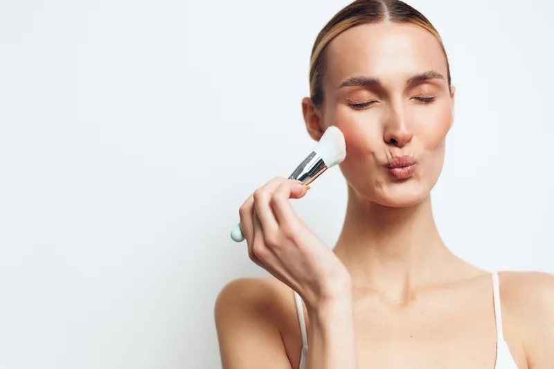 Young woman with clear skin applying makeup brush to face, eyes closed, soft natural light, minimalistic white background, beauty routine, skincare concept