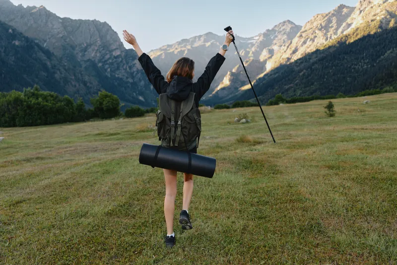 A lone hiker with a backpack raises arms in triumph across a wide meadow, with rugged mountains and a clear sky forming a dramatic natural backdrop.