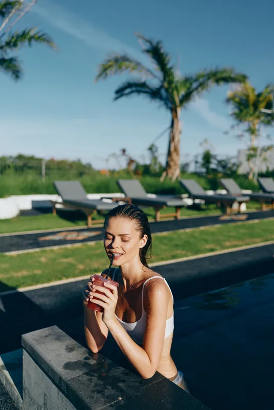 Young woman in white swimsuit relaxing in outdoor pool holding a red drink with palm trees and lounge chairs in the background on a sunny day lifestyle concept.