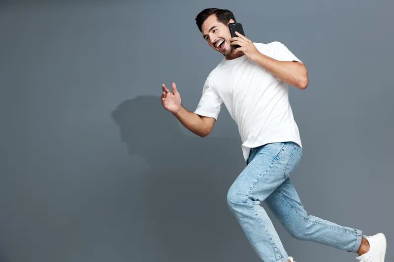 Happy young man running and talking on mobile phone, wearing a casual white t-shirt and light blue jeans, energetic expression against gray background.