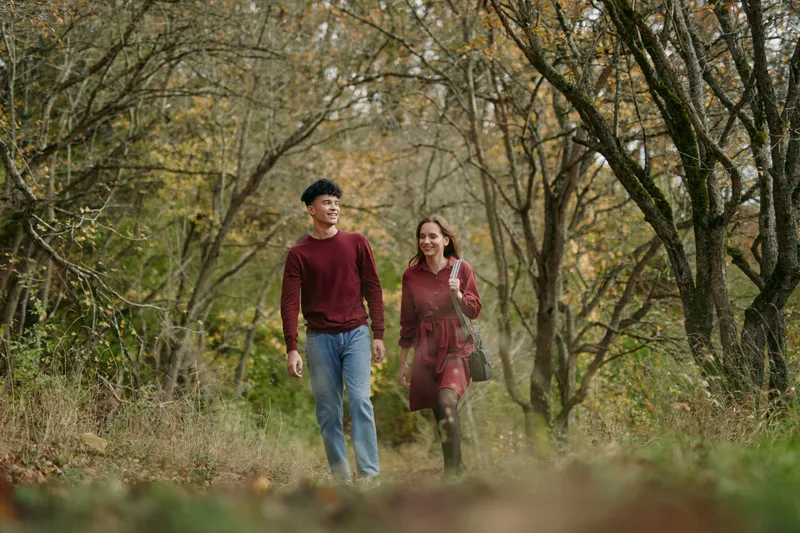 A young couple walking through an autumn woodland path, captured with natural light and candid gestures conveying authenticity and genuine connection in a relaxed everyday outdoor moment.