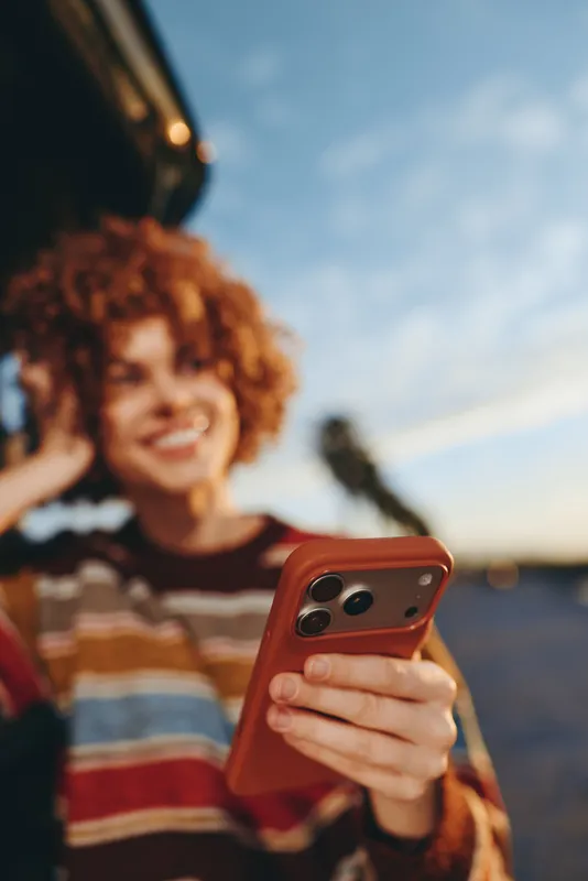 Woman outdoors, curly hair, smiling, holding smartphone close to face, laughing, sunlit warm hour light, casual striped sweater, soft blurred landscape background, lifestyle moment, natural colors