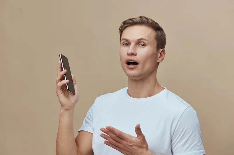 Man talking on smartphone with surprised expression, casual light blue t-shirt, indoor background, studio portrait, human emotion, communication, lifestyle concept, technology.