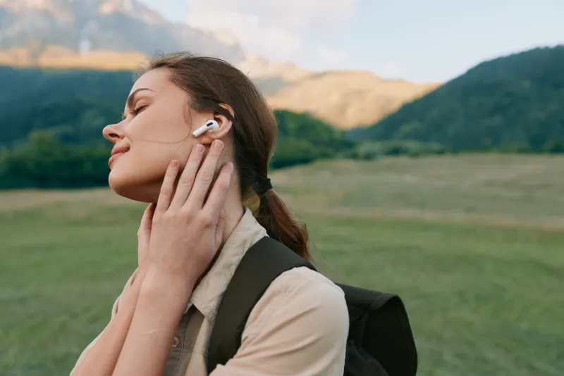 A woman outdoor headphones listening backpack casual lifestyle in nature, a landscape with mountain scenery, field and sky, sunlight and breeze creating a relaxed, comfortable, serene moment.