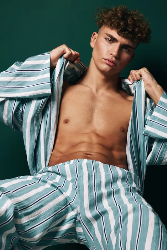 Young man with curly hair and serious look wearing striped open shirt and matching pants posing against dark backdrop showing toned chest and abs in fashion style portrait.