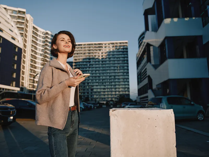 A cheerful young woman stands outdoors in a modern urban setting, texting on her phone. She smiles confidently, suggesting productivity, optimism, and a dynamic, forward moving workday.
