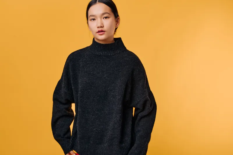 Young woman wearing black sweater standing against yellow background with calm expression and straight posture posing for fashion portrait studio image.