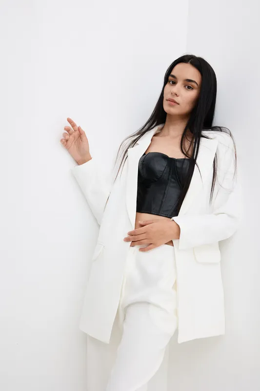 Young woman wearing white suit and black top poses confidently against white wall, showcasing stylish fashion and modern elegance with natural expression.