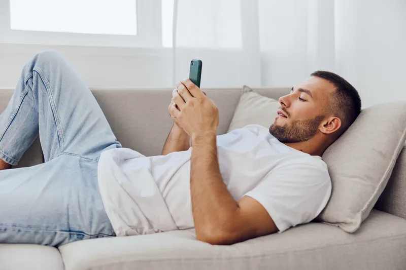 Relaxed man using smartphone on a cozy sofa, showcasing modern lifestyle and technology. Bright natural light enhances the calm atmosphere in the room.