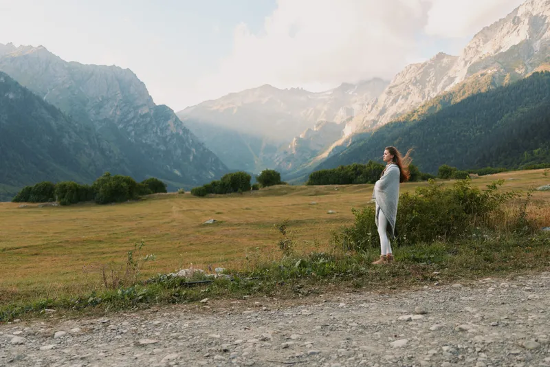 A woman in casual clothes stands on a dirt path in a vast mountain valley surrounded by green trees and rocky peaks under a cloudy sky. Lifestyle concept outdoors.