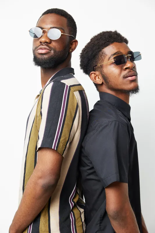 Confident black men wearing stylish sunglasses stand back-to-back, showcasing their unique fashion sense and modern vibe in a bright studio setting.