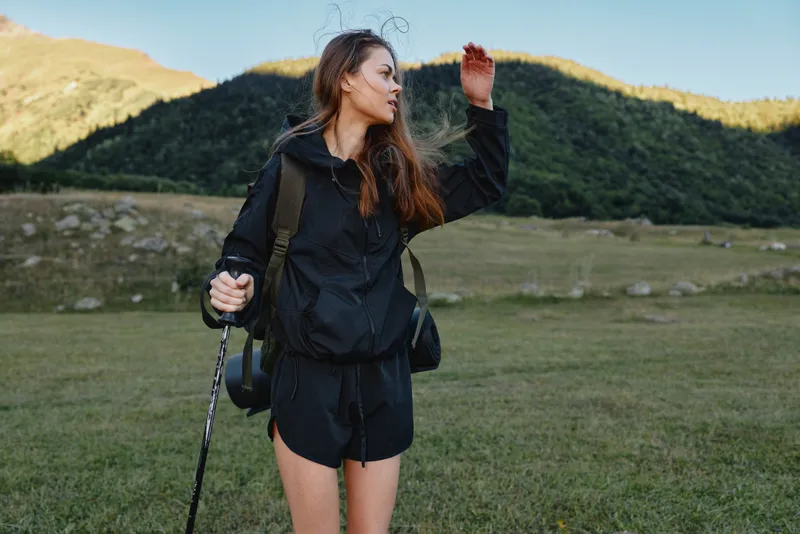 A young woman hikes through a wide meadow with a backpack, wearing a dark outfit. She gazes into the distance with a calm, thoughtful smile, enjoying nature and a peaceful outdoor adventure.