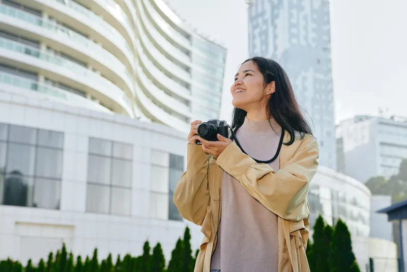 Photography of a woman with a camera in an urban outdoor setting, looking up at modern buildings, capturing cityscape and street life, showcasing creativity and curiosity in a travel mood