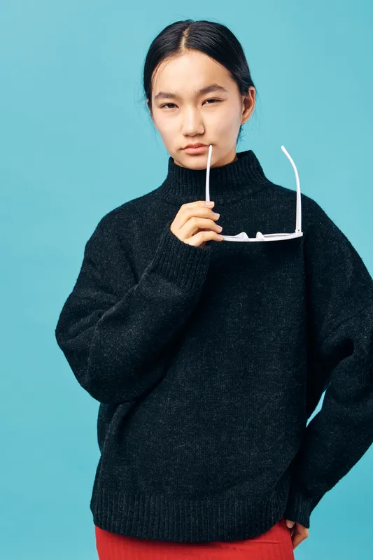 Young woman with black hair and black turtleneck sweater holding white eyeglasses on blue background. Casual style, serious look and minimalist fashion portrait for modern lifestyle.