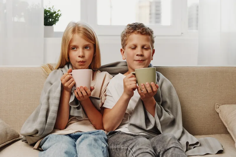 Two cozy children wrapped in blankets enjoy warm drinks sitting on a sofa in bright natural light at home.