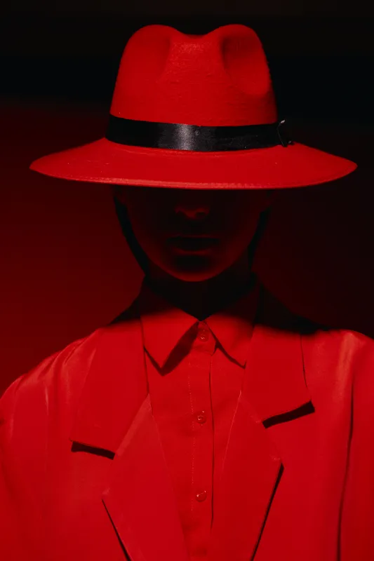 Mysterious woman in a striking red hat and jacket stands in low light, emphasizing shadows across her face. Creative fashion concept with bold color contrast and style.