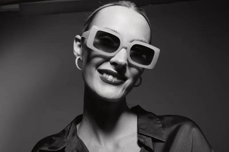 Stylish white woman with pale skin wearing oversized sunglasses and earrings smiling in black and white close-up.