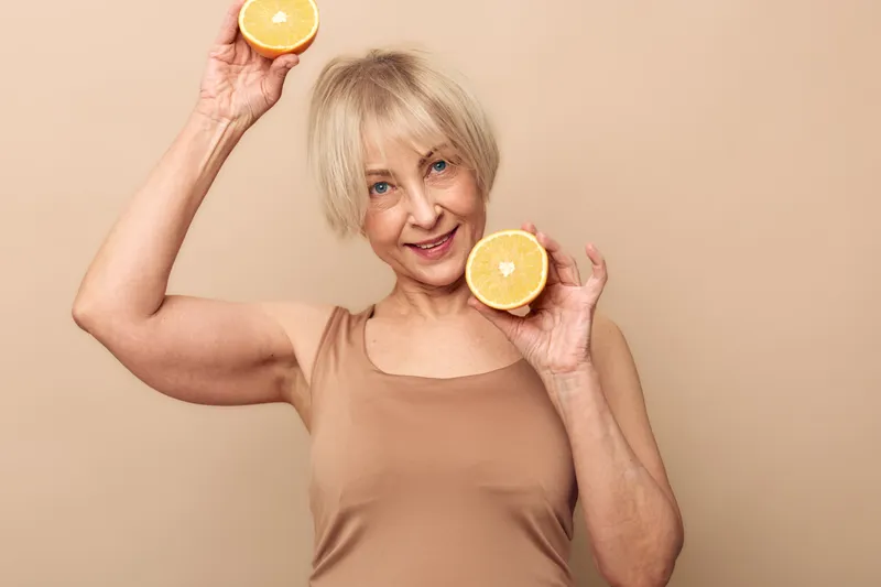 Smiling middle aged woman with short blonde hair holds two halves of fresh orange near face and raised hand, wearing sleeveless top on beige background. Concept of healthy lifestyle and vitamin C.