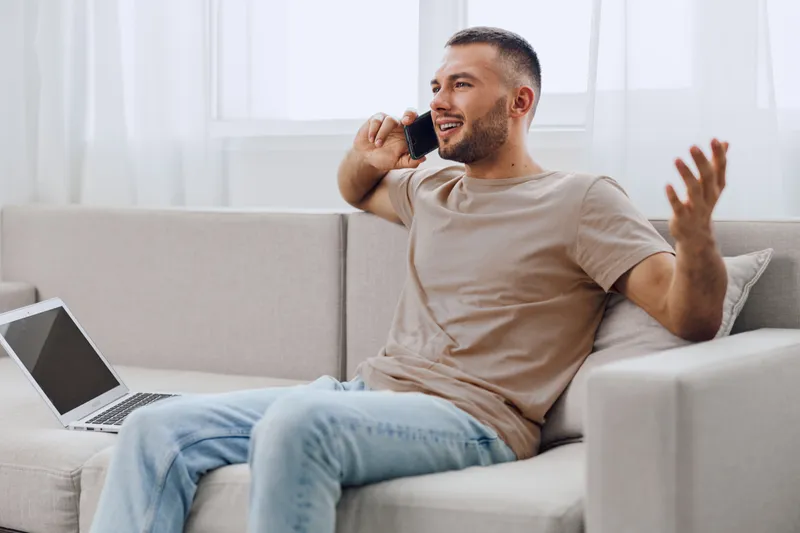 Smiling man engaged in a lively phone conversation while sitting on a modern sofa, dressed casually and surrounded by a bright, airy interior, conveying energy and positivity.