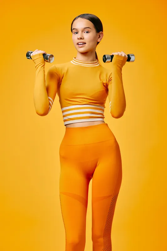 Fit woman in orange athletic wear lifts dumbbells against a vibrant yellow backdrop, showcasing strength, balance and focus during a dynamic workout session.