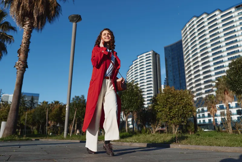 Young woman in a red coat walks confidently along a city sidewalk on a sunny day with modern buildings and palm trees in the background.