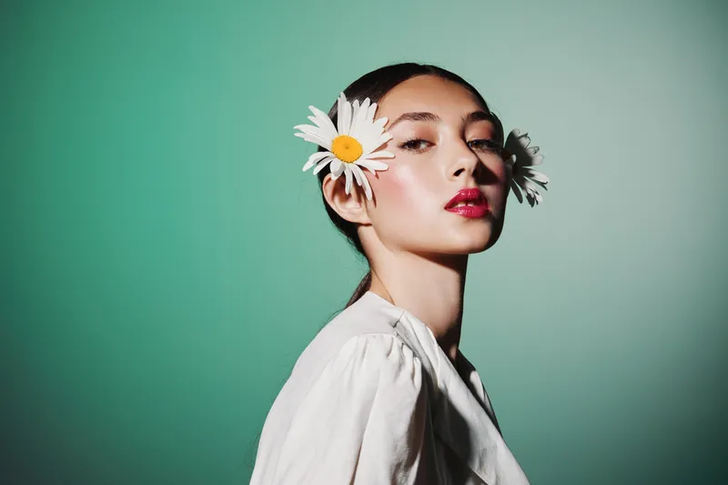 Young woman with bright makeup and distinctive red lipstick poses with two large white daisies in her hair on a turquoise background. Fashion and beauty creative concept.