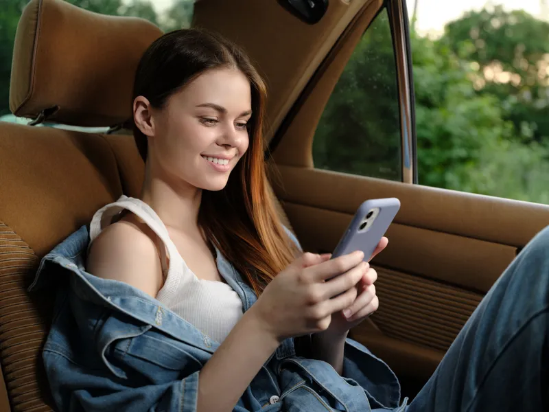 Young woman smiling while using smartphone in a car, dressed in casual denim outfit, with a green nature background, evoking feelings of joy and freedom.