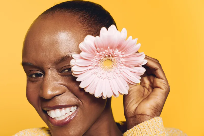Smiling black woman with dark skin holds pink flower near eye on yellow background. Close-up portrait of cheerful African female model with natural beauty and positive expression.