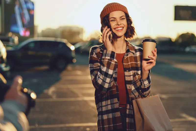 Woman holding coffee and shopping bag, smiling on phone in parking lot at sunset, candid lifestyle portrait with authenticity, golden hour glow, mindful living and emotional storytelling.
