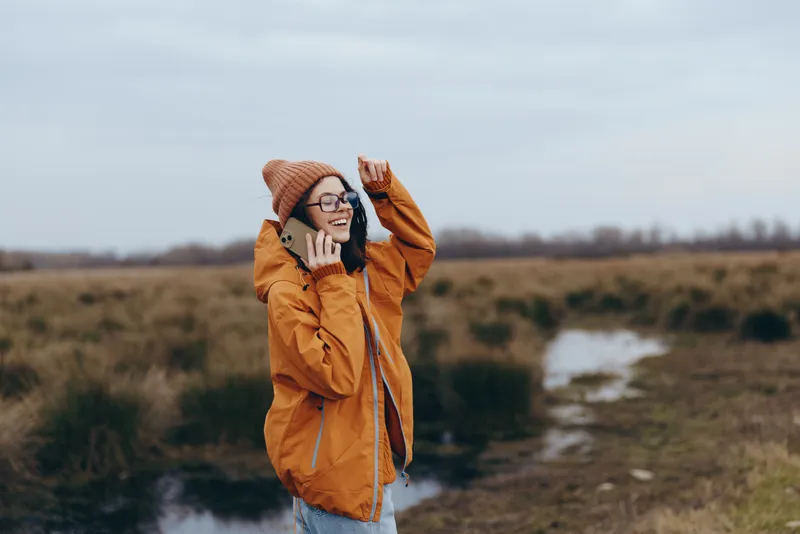 Woman lifestyle outdoors in a field, wearing warm hat and glasses, adjusting her hat with both hands, listening to headphones, autumn nature under cloudy sky, candid portrait today